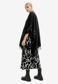 Poncho nero con bordi frangiati, caratterizzato da un motivo audace. Abbinato a una lunga gonna astratta stampata in bianco e nero e stivali neri con suola spessa.