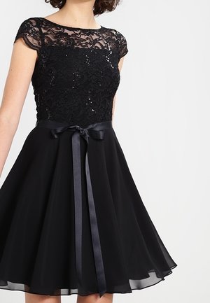 Mujer con un vestido negro de encaje con mangas cortas, una cinta de satén atada a la cintura y una falda de gasa acampanada hasta la rodilla.