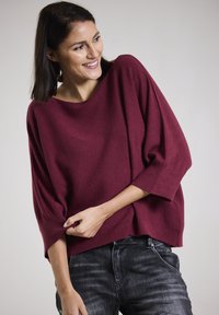 Burgunderfarbener Oversized-Pullover mit weitem Ausschnitt und Dreiviertelärmeln, aus einem weichen Stoff, kombiniert mit dunklen Jeans.