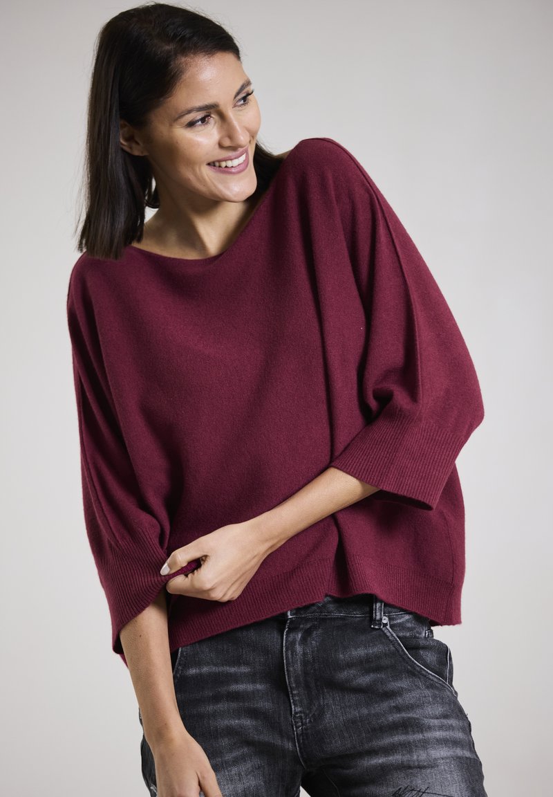 Burgunderfarbener Oversized-Pullover mit weitem Ausschnitt und Dreiviertelärmeln, aus einem weichen Stoff, kombiniert mit dunklen Jeans.