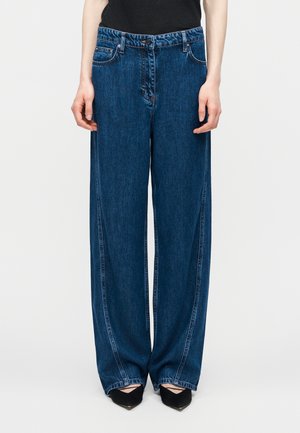 Γυναίκα που φοράει άνετο σκούρο μπλε τζιν και μαύρα παπούτσια με μυτερή μύτη, στέκεται μπροστά σε απλό φόντο.