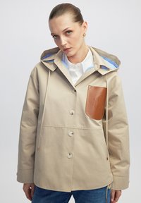 Beżowa wodoodporna kurtka z kapturem, z brązową skórzaną kieszenią, zapięciem na guziki i niebieską w paski podszewką. Gładka tekstura, casualowy design.