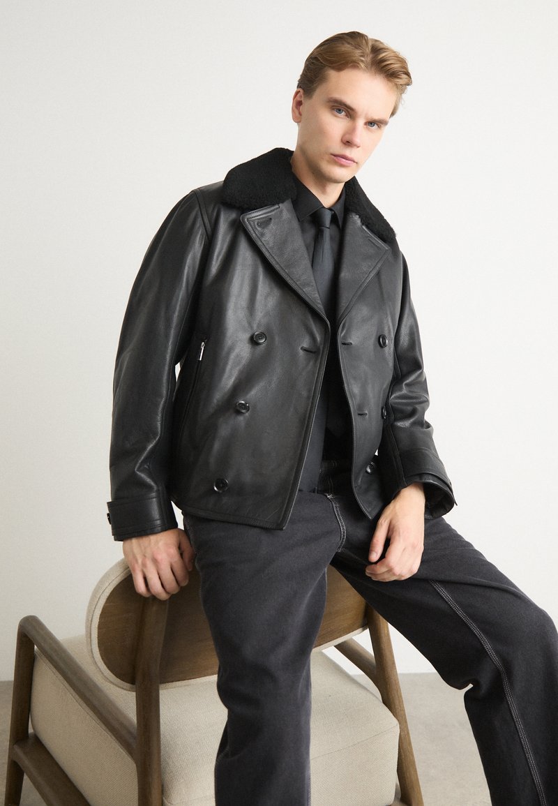 Mustat nahkainen kaksirivinen takki, jossa on pehmeä musta kaulus, vetoketjulliset taskut ja sileä pinta. Mallilla on päällään tummat farkut.