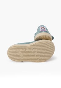 Hellgrüne Leinenschuhe mit beige Gummisohle, ausgestattet mit einem Etikett im Cartoon-Design. Die Sohle hat horizontale und vertikale Rutschmuster.