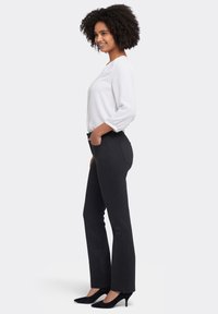 Svarta skräddarsydda byxor i ett texturerat tyg, kombinerade med en vit långärmad blus. Modellen bär svarta högklackade skor.