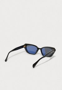 Sorte rektangulære solbriller med blåtonede linser som hviler på en hvit overflate, og viser detaljer fra innersiden av hengslet og armen.