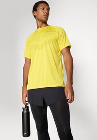 Camiseta deportiva amarilla con un patrón texturizado, combinada con pantalones cortos negros y mallas. Se sostiene una botella de agua negra en la mano derecha.