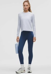 Világoskék, hosszú ujjú sport felső, tengerészkék leggings, szürke sportcipők. A modell egy világoskék kulacsot tart, amely elegáns és testhezálló dizájnnal rendelkezik.