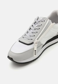 Harmaa urheilukenkä, jossa valkoista verkkoa ja tekonahkapaneelit, varustettu litteillä nauhoilla, vetoketjidetaljilla sekä mustalla kumipohjalla, jossa kohokuvioitu brändäys.