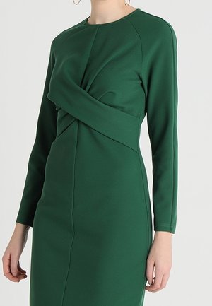 Femme portant une robe à manches longues vert foncé avec un détail en tissu torsadé sur la poitrine et la taille.