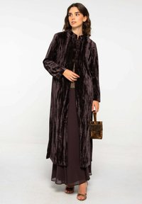 Long manteau en velours violet foncé avec un col haut et une fermeture à toggles à l'avant. Associé à une robe opaque à couches et un sac à main scintillant.