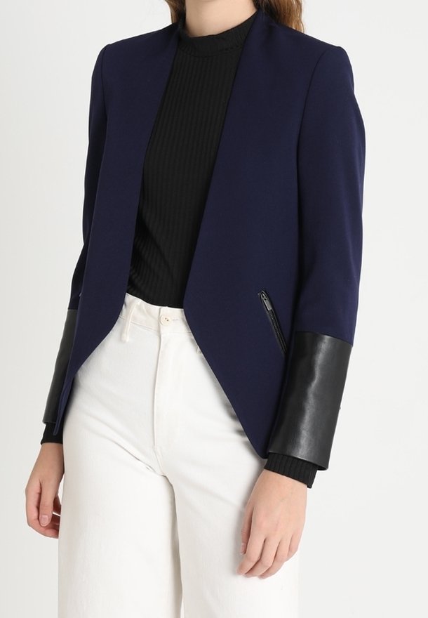 Blazer marine à la coupe structurée, avec des manches en cuir noir et des poches zippées. Porté sur un haut noir à côtes et un pantalon blanc.