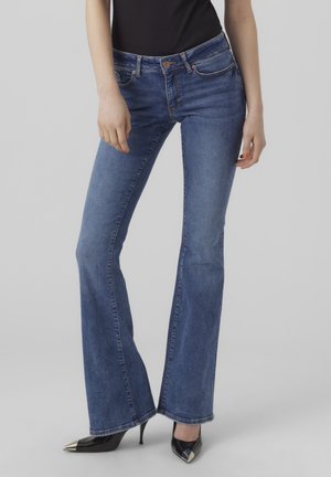 Persoon die mid-rise blauwe denim flared jeans draagt met zwarte, puntige hoge hakken en een zwart mouwloos topje, staand tegen een effen achtergrond.