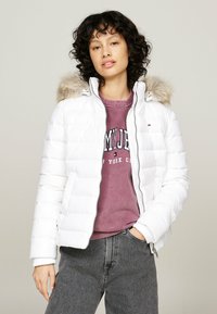 Donna con capelli ricci e scuri, che indossa una giacca imbottita bianca con cappuccio di pelliccia, una felpa viola e jeans grigi, in piedi con una mano in tasca.