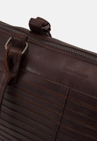 Czarna, teksturowana torba ze skóry o ciemnym odcieniu, z metalowym suwak i metalowymi pierścieniami, z wytłoczonym logo marki na przednim panelu.