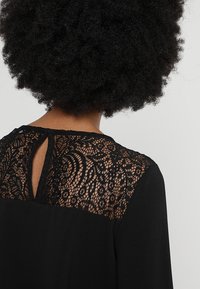 Svart blus med spetsbakgrund i intrikata swirlmönster och en liten nyckelhålsöppning. Slätt tyg med en avslappnad passform.