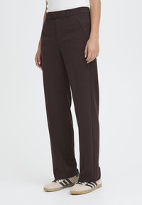Pantalons amples marron avec une texture lisse, dotés de poches latérales et d'une ceinture, associés à des baskets légères avec des rayures noires.