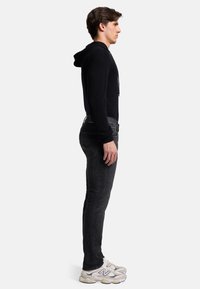 Schwarzer Kapuzenpullover, taillierte Passform. Schwarze Jeans, slim fit. Weiße und graue Sneaker mit Logo und strukturierten Sohlen. Seitenansicht.