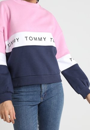 Osoba ubrana w różową, białą i granatową bluzę Tommy z wyszywanym logo, w połączeniu z niebieskimi jeansami, stojąca na jednolitym tle.