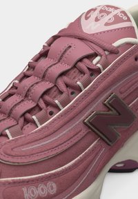 Κοντινό πλάνο από ροζ αθλητικό παπούτσι New Balance 1000 με σουέντ υφή, ραμμένες λεπτομέρειες και το εμπορικό σήμα "N" στο πλάι.