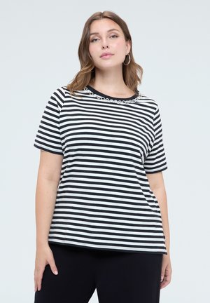 Vrouw draagt een zwart-wit horizontaal gestreept T-shirt met korte mouwen en kettingdetail bij de halslijn en zwarte broek, staand tegen een eenvoudige achtergrond.