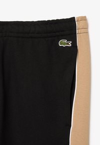 Pantalons de sport noirs avec un panneau latéral beige, des finitions blanches et un logo crocodile brodé vert sur le côté gauche.