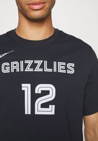 Černý atletický dres s jemnou texturou obsahuje výrazné bílé písmo s nápisem "GRIZZLIES" a číslem "12". Na rameni je logo Nike v duhovém designu.