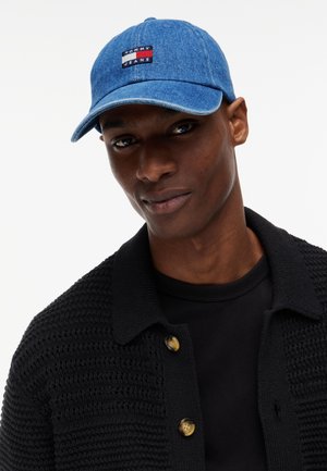Ung mand iført en blå denim Tommy Jeans kasket og en sort strikket jakke med store knapper over en sort skjorte, der vender fremad.