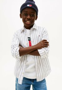 Striped, collared, button-up shirt in white and blue, worn over a graphic t-shirt. Dark knit beanie with logo. Jeans with a relaxed fit.