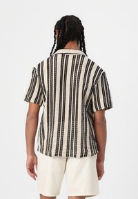 Camisa de manga corta en crema con rayas verticales negras. Presenta un cuello con solapa y una tela texturizada, con un ajuste relajado.