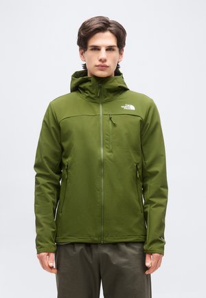 Νεαρός άνδρας που φοράει μπουφάν North Face σε ελιά πράσινο χρώμα με μπροστινό φερμουάρ και κουκούλα, στέκεται μπροστά σε απλό φωτεινό φόντο.