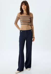 Maglione a coste color tan con righe navy, abbinato a pantaloni a gamba larga navy con dettagli in oro e grandi tasche. Vestibilità casual.