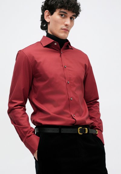 Crvena košulja na dugih rukava s širokim ovratnikom i tamnim gumbima, uparena s crnim remenom i tamnim hlačama. Glatka tkanina.