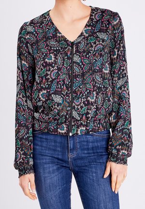Femme portant un chemisier noir à fermeture éclair avec un motif floral multicolore et un jean bleu, debout devant un fond uni.
