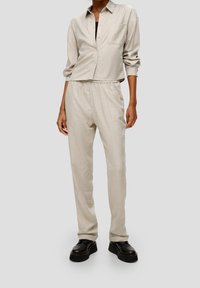 Beige, relaxed-fit button-up shirt with single chest pocket, paired with matching elastic-waist trousers. Black chunky-soled shoes.