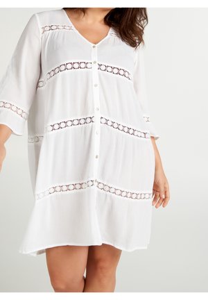 Woman wearing a loose white button-down dress with three-quarter sleeves and horizontal lace panels across chest, sleeves, and skirt.