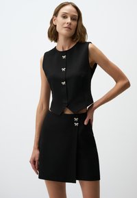 Completo due pezzi nero composto da un top senza maniche con dettagli a fiocco argento e una gonna coordinata con un design avvolgente diagonale.