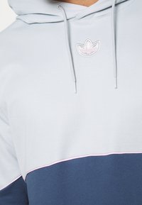 Světle modrá mikina s kapucí s tmavě modrým spodním panelem, která má růžovou akcentovou linku a vyšitý logotyp uprostřed.