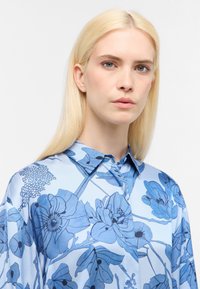 Camicia di seta azzurra chiara con stampa floreale in varie sfumature di blu. La camicia presenta un colletto a punta e una fila di bottoni.