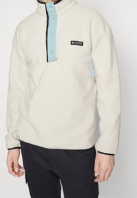 Pullover in pile color crema con patta blu a scatto e tasca con zip. Presenta rifiniture nere e un tessuto morbido e texturizzato.