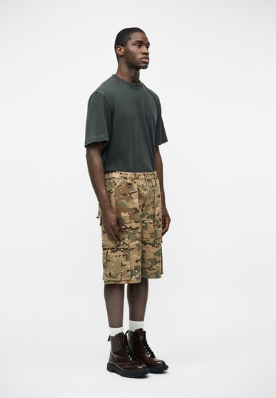 Νεαρός άντρας που στέκεται, φοράει γκρι σκουρόχρωμο φαρδύ T-shirt, παραλλαγής cargo σορτς, λευκές κάλτσες και σκούρα καφέ μποτάκια με κορδόνια, σε λευκό φόντο.