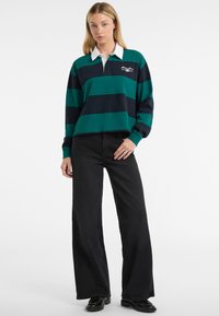 Polo a maniche lunghe a righe verdi e blu con colletto bianco e logo ricamato, abbinato a jeans neri a vita alta e ampia e scarpe.