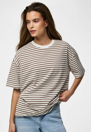 Kvinde med langt brunt hår, iført en hvid og brunstribet t-shirt og lyseblå jeans, trækker let i trøjen til siden.