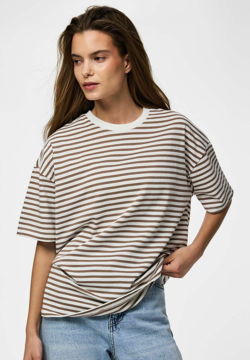 Kvinne med langt brunt hår iført en hvit og brunstripet t-skjorte og lyseblå jeans, trekker skjorten litt til siden.