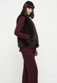 Gilet en fausse fourrure marron foncé, offrant une couverture complète à l'avant et une finition douce et texturée. Porté sur une tenue bordeaux profonde au fit décontracté.
