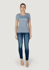Vaaleansininen istuva t-paita, jossa on kiiltävä "GUESS LOS ANGELES" -logo, yhdistettynä tummiin skinny-farkkuihin ja nudet kärkitossut.