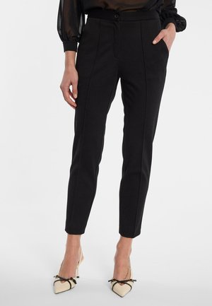 Vrouw draagt zwarte, op maat gemaakte broek tot op de enkel met naden aan de voorkant en beige, puntige hoge hakken met zwarte strikken.