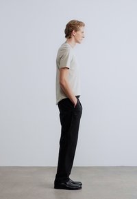 Μπεζ κοντομάνικο μπλουζάκι και μαύρο παντελόνι σε μοντέλο. Το μπλουζάκι είναι μονόχρωμο με άνετη εφαρμογή, το παντελόνι έχει ευθεία γραμμή. Μαύρα παπούτσια ολοκληρώνουν το σύνολο.