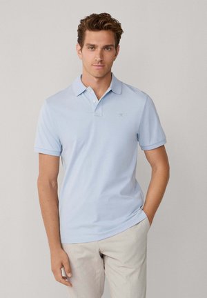 Polo azul claro hecho de tela suave con collar, mangas cortas y un bordado sutil en el pecho. Combinado con pantalones beige.