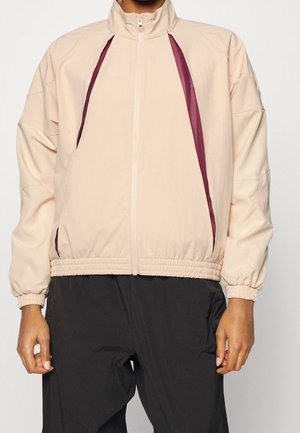 Veste beige à fermeture éclair avec des accents de rayures diagonales bordeaux, portée avec un pantalon noir, poignets élastiques et ourlet inférieur.
